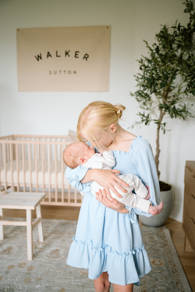 Big sister holding her newborn sibling, both bathed in natural window light.